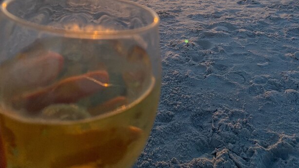 Una copa de vino en una playa arenosa con el océano y una puesta de sol al fondo.