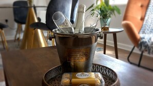 Ein Esstisch mit einem Metallkübel, der Champagnergläser und eine Flasche enthält, einer Schale mit Ferrero-Schokoladen und einer Vase mit Blumen.
