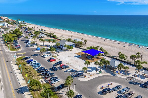 An aerial view of a coastal area with a beach, parking lot, and a building with a blue roof.