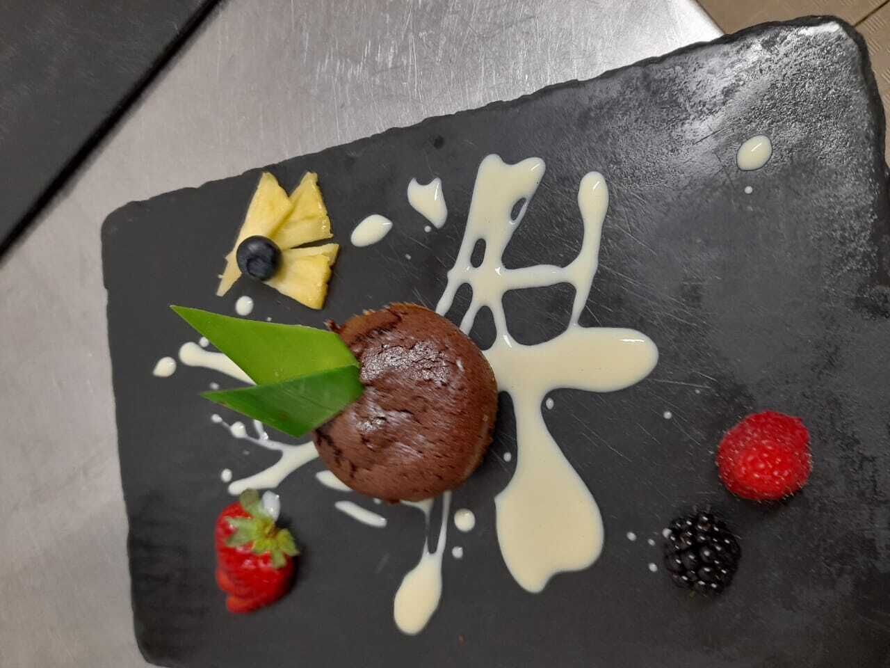 Un dessert servi sur une assiette d’ardoise noire, accompagné d’une sauce blanche, d’une fraise, d’une framboise, d’une mûre, d’une myrtille et d’une feuille verte.
