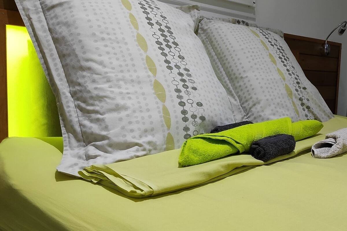 A neatly made bed with a green bedspread, pillows with a dotted pattern, and folded towels in green and grey.