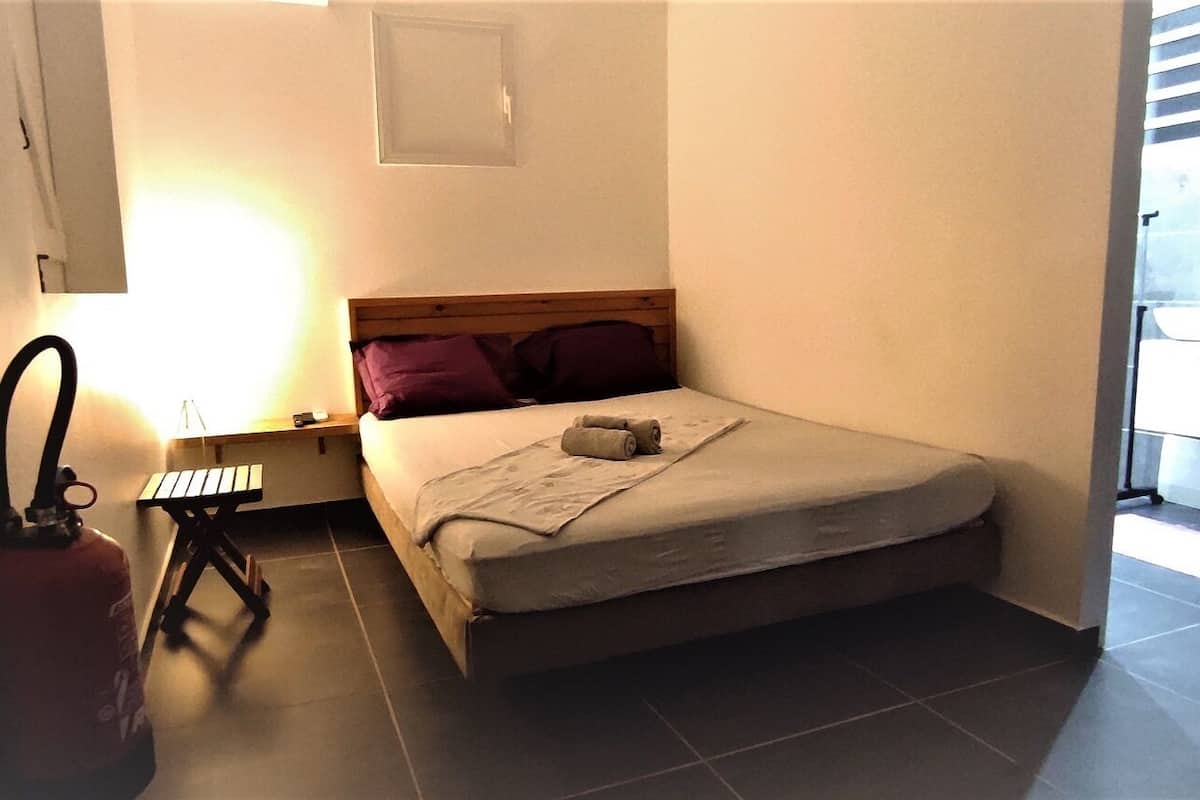 A small, compact room with a bed, a nightstand, a lamp, and a window.