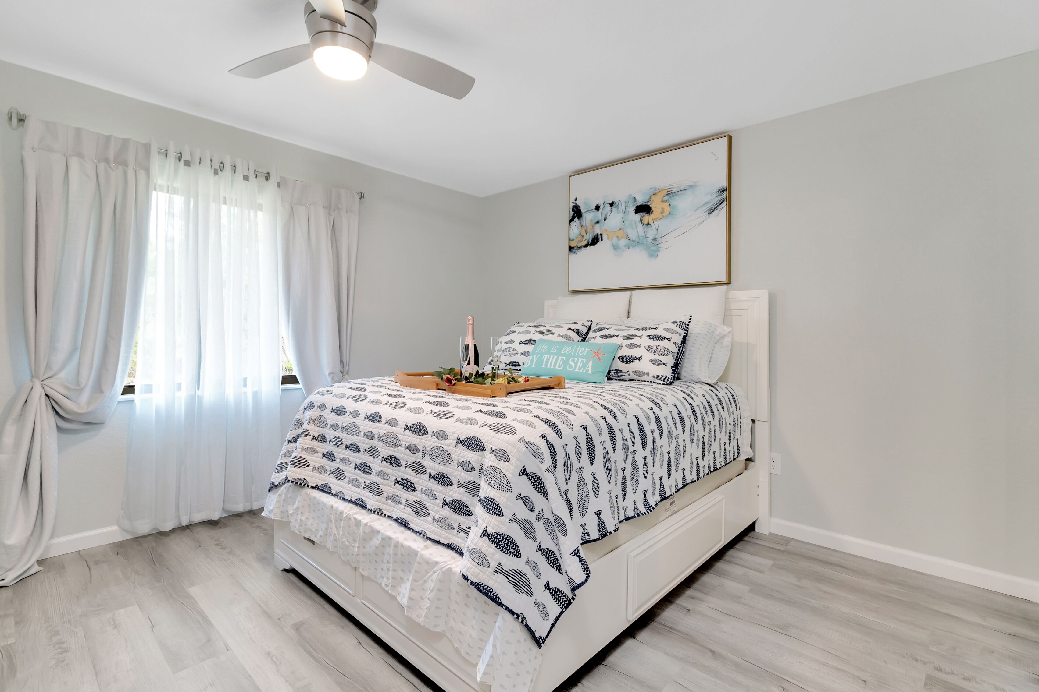 Une chambre à coucher comprenant un lit, un ventilateur de plafond, une fenêtre avec des rideaux et une œuvre d’art accrochée au mur.