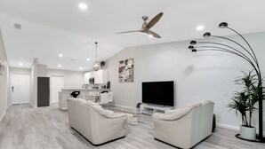 Ein modernes Wohnzimmer mit einem Flachbildfernseher, zwei Sofas, einem Deckenventilator und einem Essbereich mit einer Küche im Hintergrund.