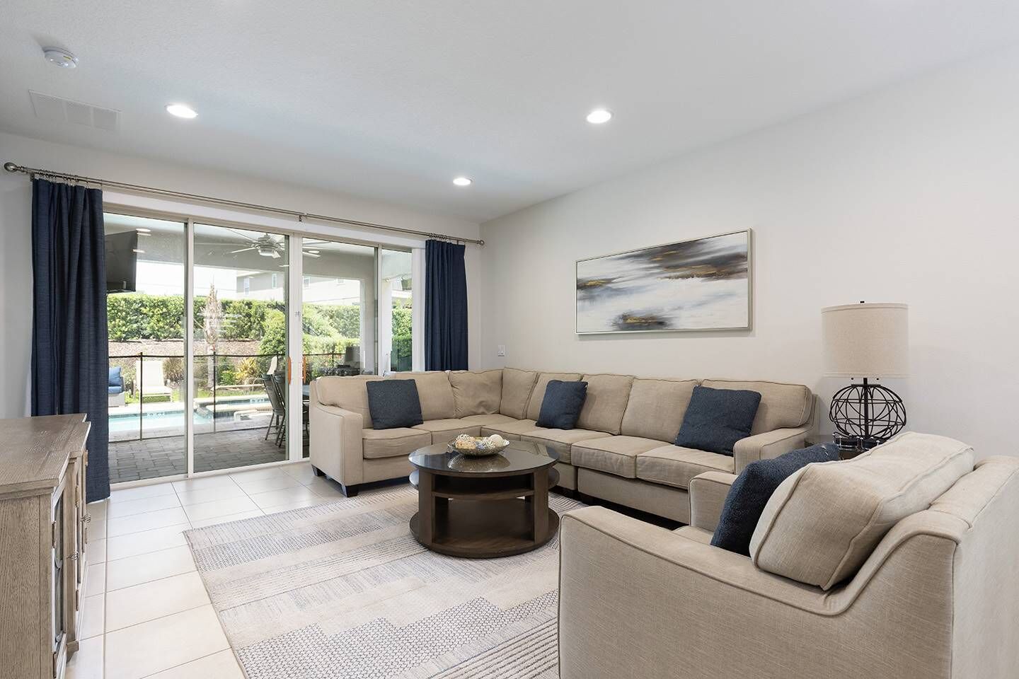 Un salon moderne avec un canapé d’angle beige, une table basse et une lampe. Une porte-fenêtre coulissante donne accès à un balcon avec une piscine.