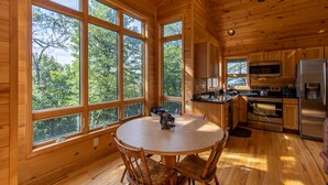 Una cabaña de madera con una mesa redonda y sillas, ventanas amplias, una cocina con electrodomésticos modernos y vistas al bosque.