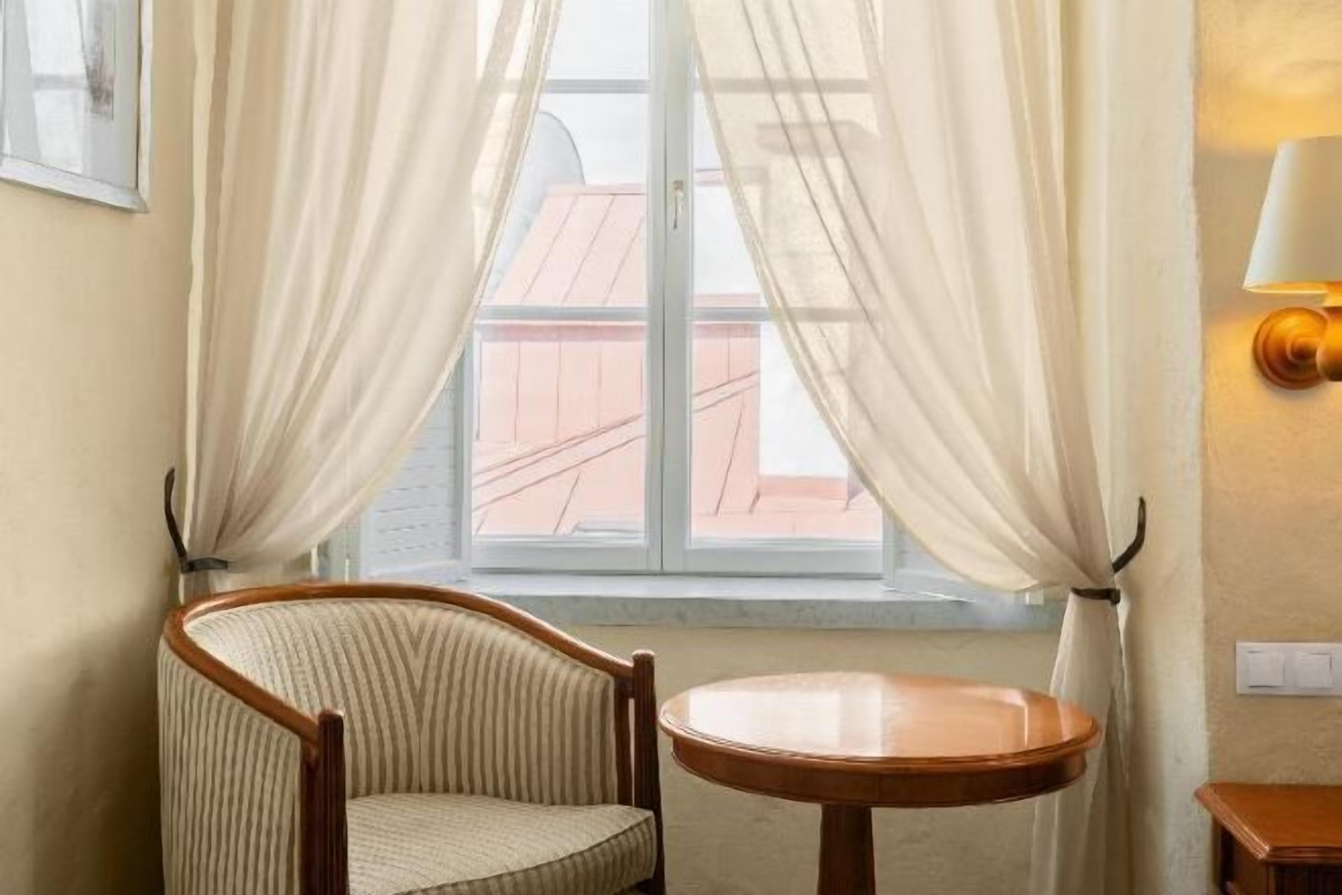 A room with a chair, a small round table, sheer curtains, a window with a view of buildings, and a wall-mounted lamp.