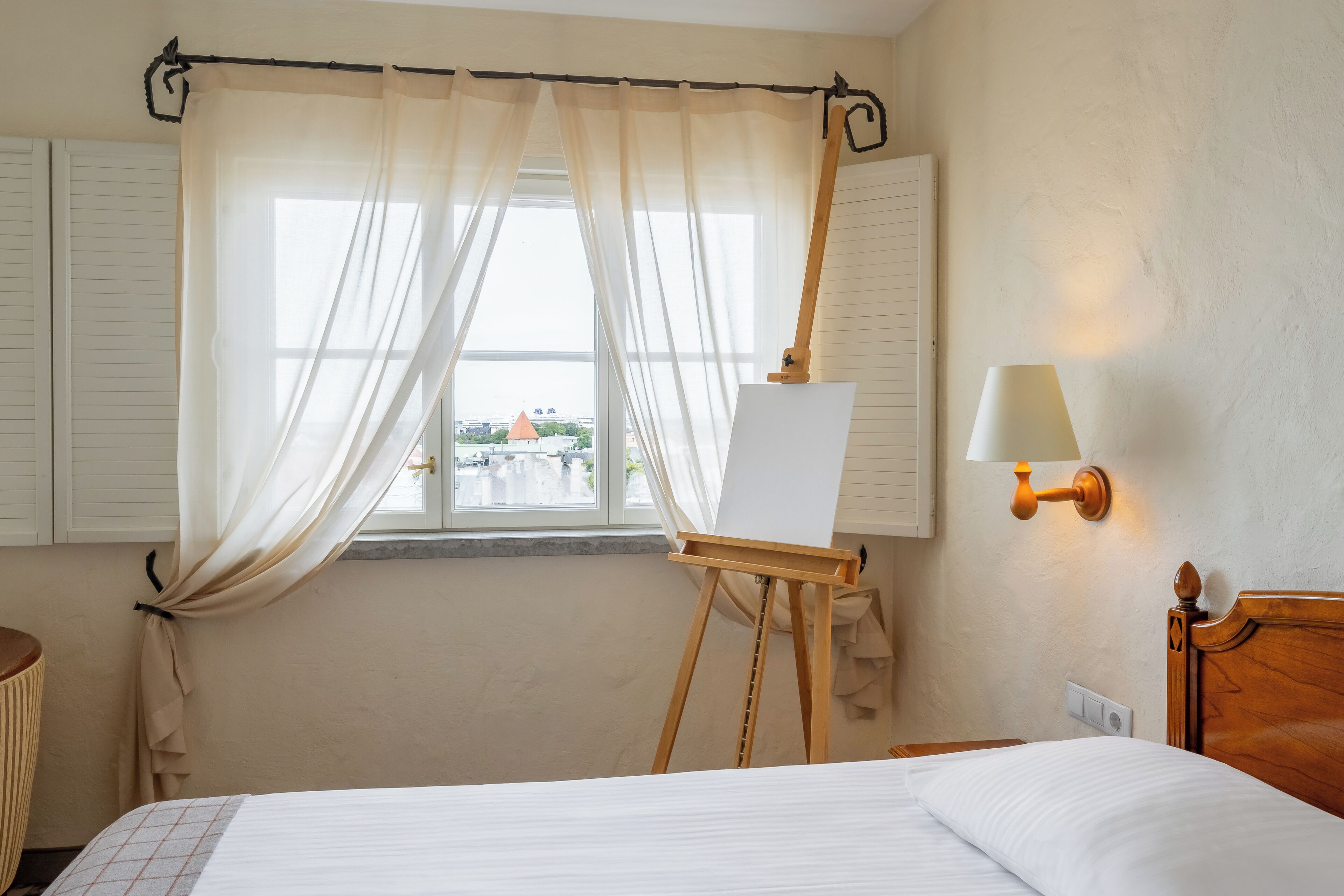A hotel room with a bed, a window with curtains, an easel with a blank canvas, and a wall-mounted lamp.