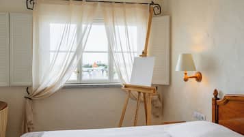 A hotel room with a bed, a window with curtains, an easel with a blank canvas, and a wall-mounted lamp.