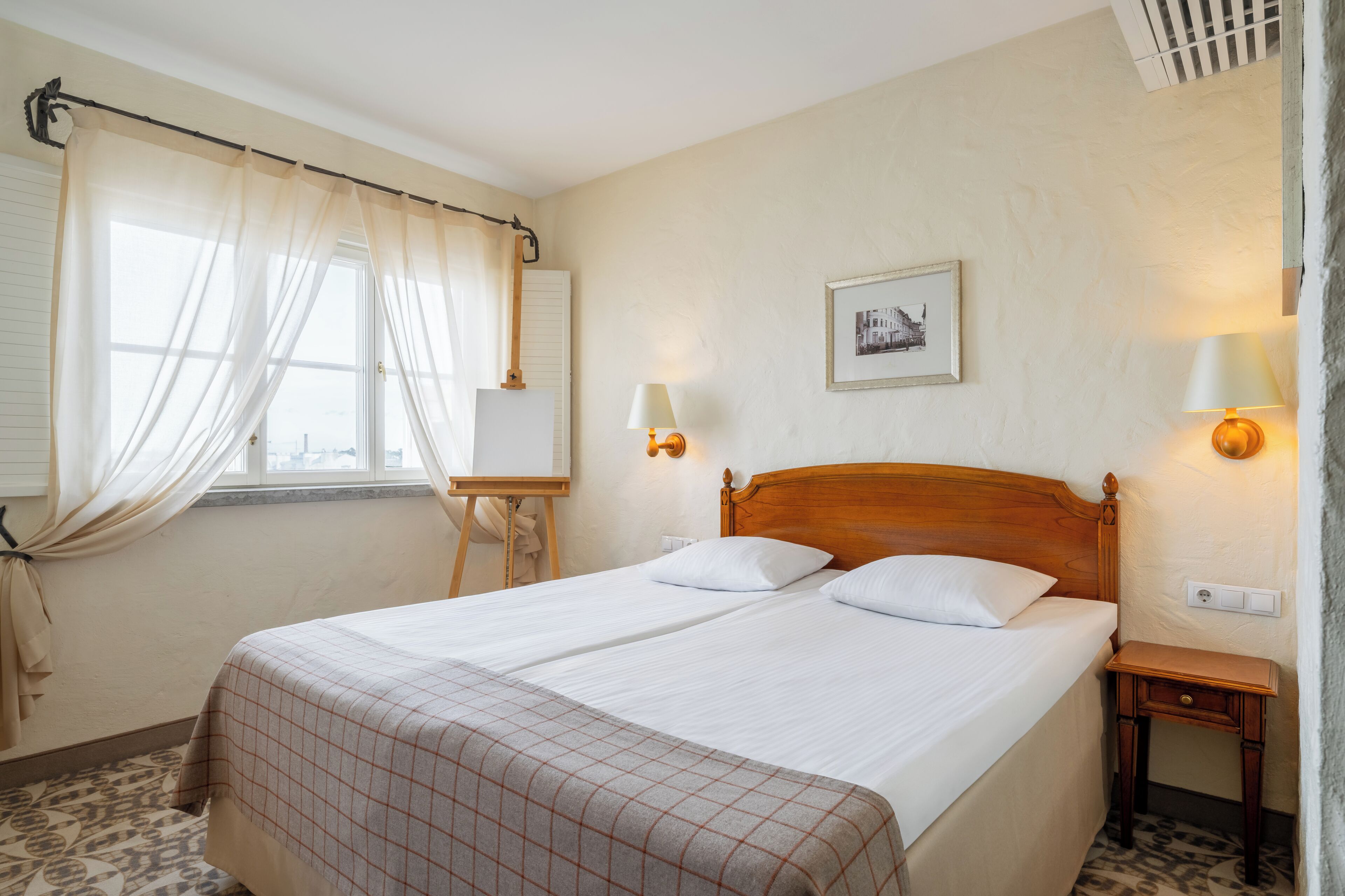 Una cama doble con cabecera de madera, una mesita de noche, una lámpara y una ventana con cortinas traslúcidas.
