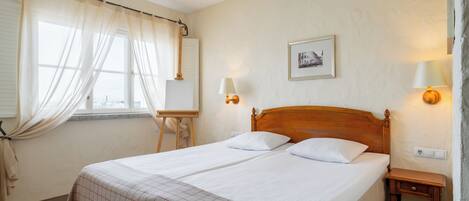 Una cama doble con cabecera de madera, una mesita de noche, una lámpara y una ventana con cortinas traslúcidas.