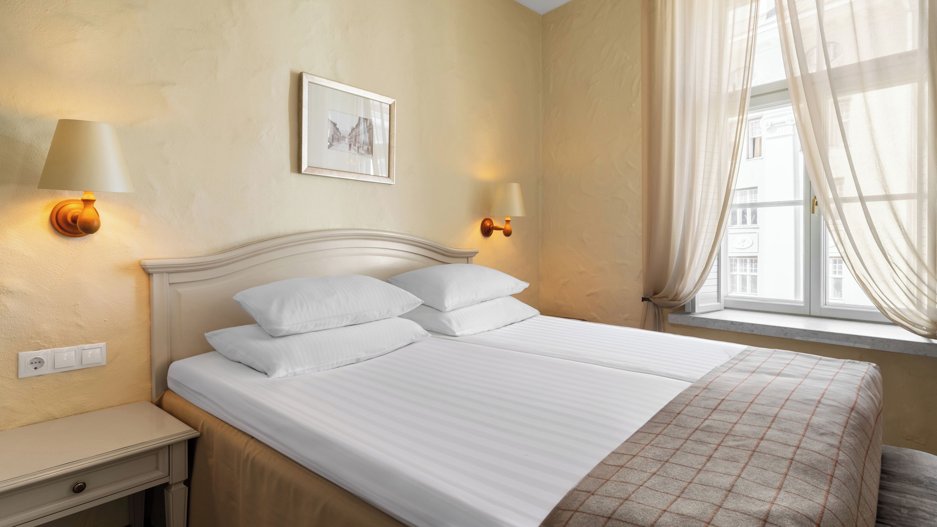 A neatly made bed with white linens, a bedside table with a lamp, and a window with sheer curtains.