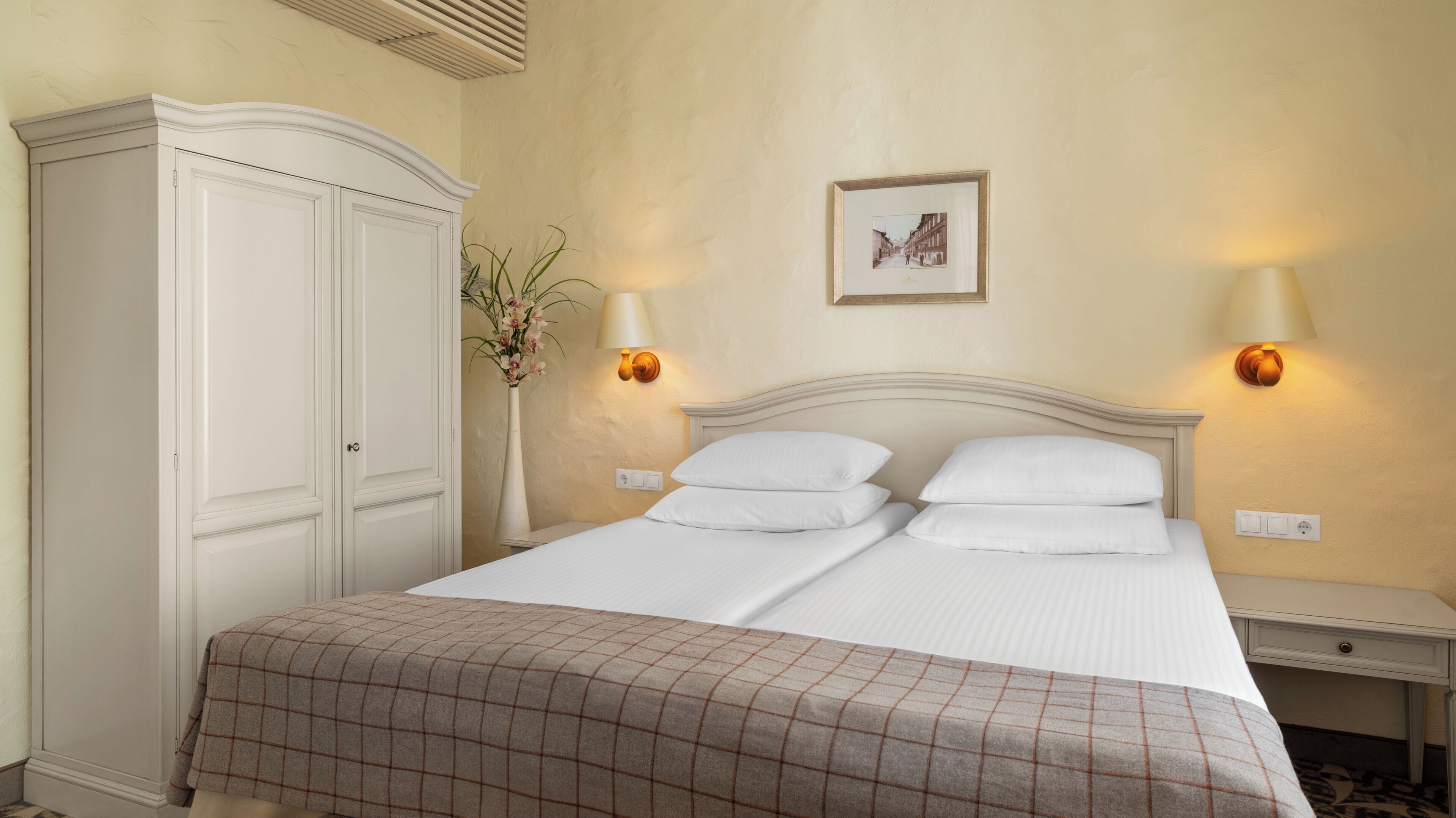 Una cama bien hecha con sábanas blancas y una manta escocesa, un armario blanco y dos lámparas de pared.