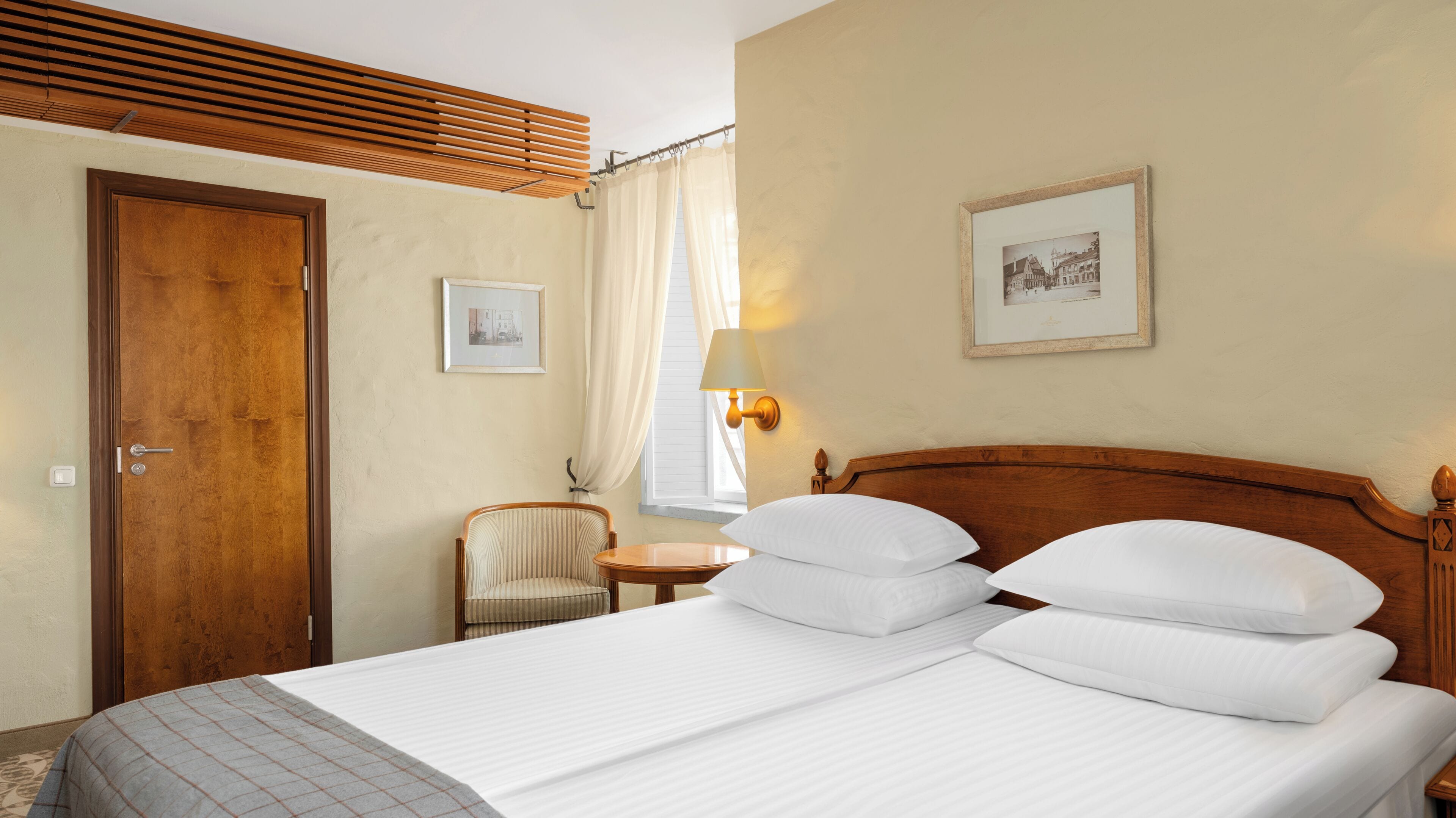 A hotel room with a wooden headboard, a bed with white linens, a small bedside table with a lamp, a chair, and a framed picture on the wall.