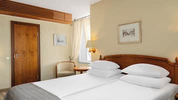 A hotel room with a wooden headboard, a bed with white linens, a small bedside table with a lamp, a chair, and a framed picture on the wall.
