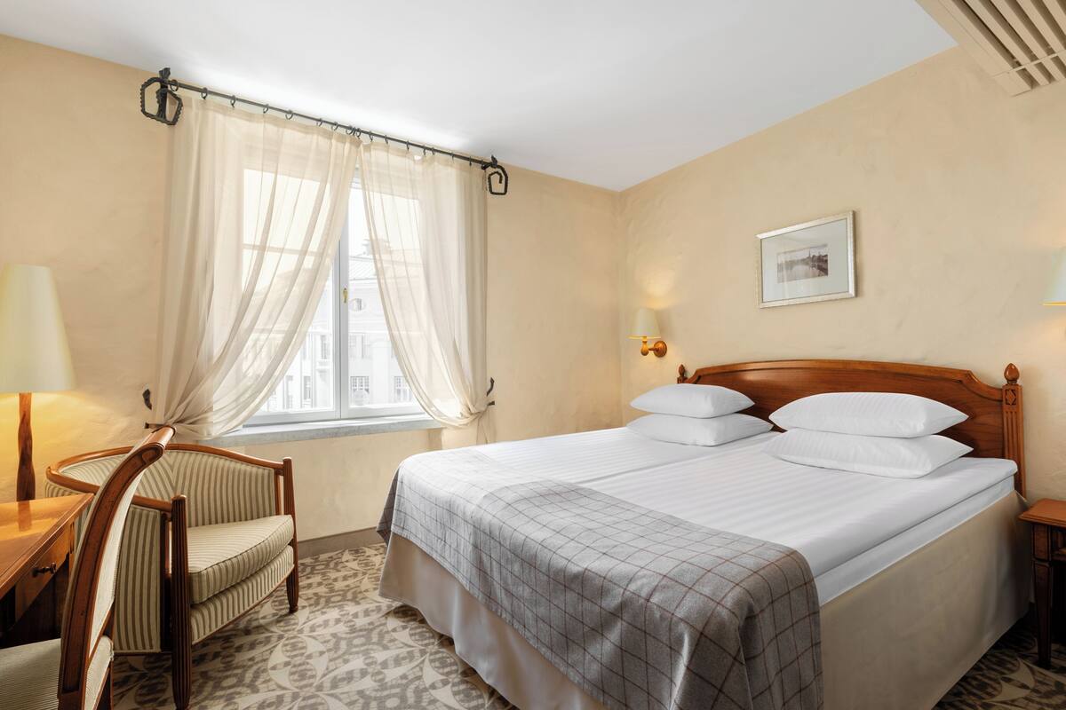 Une chambre d’hôtel comprenant un lit, un bureau, une chaise, une fenêtre avec des rideaux légers, un tableau accroché au mur et deux lampes de chevet.