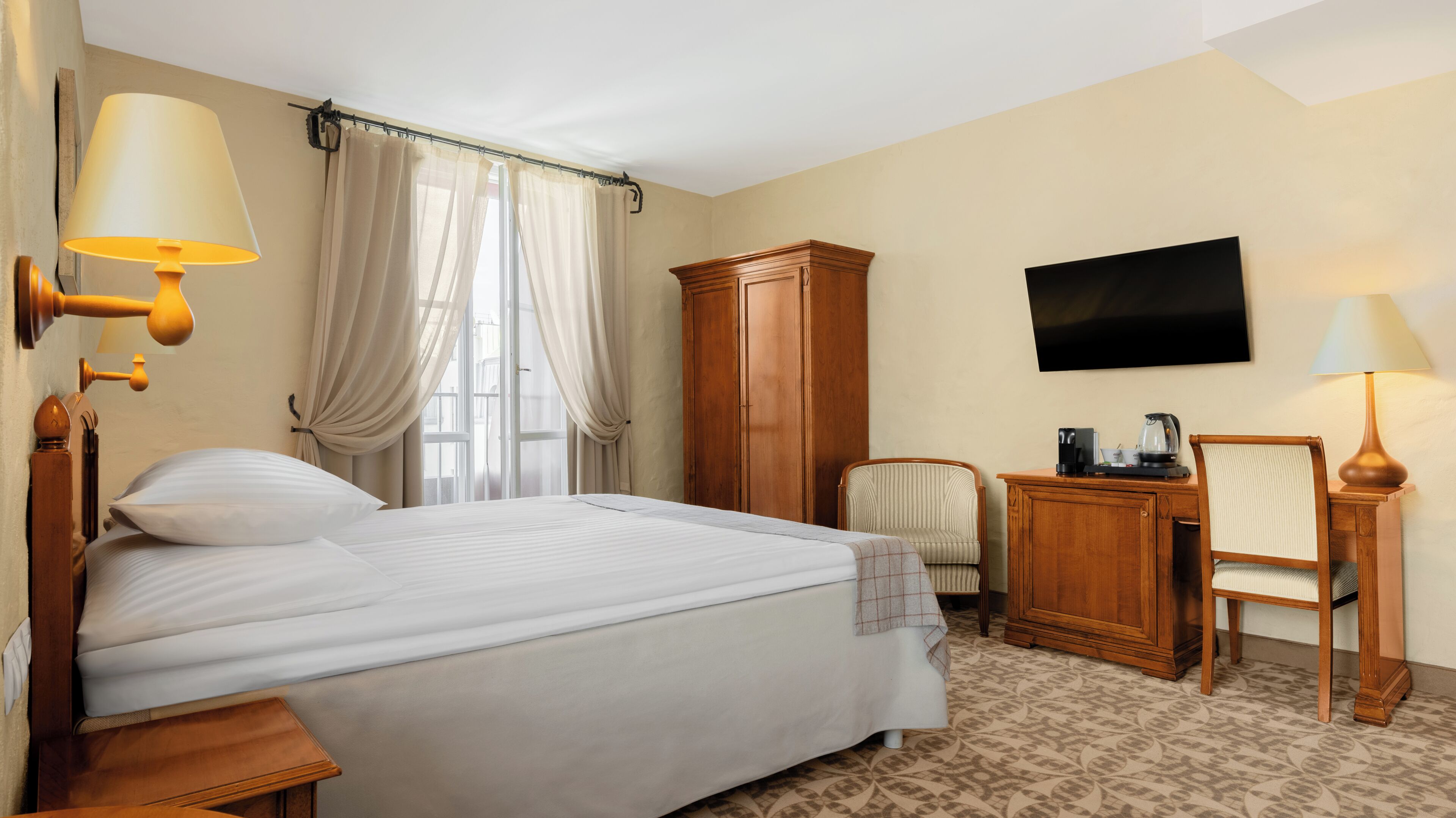 Une chambre d’hôtel comprenant un lit, une armoire en bois, une télévision, un bureau avec une chaise et une fenêtre avec des rideaux.