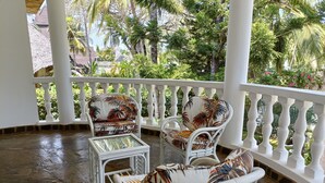 Uma área de estar coberta ao ar livre, com móveis de vime branco e uma mesa de vidro, cercada por um parapeito branco e vegetação tropical exuberante.