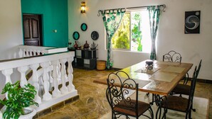 Uma área de refeições com mesa de madeira, cadeiras de metal preto, balaústres brancos e vista para o verde através de uma janela com cortinas estampadas.
