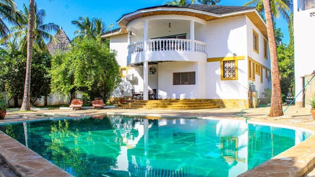 Uma casa de dois andares com piscina, cercada por palmeiras e um céu azul límpido.