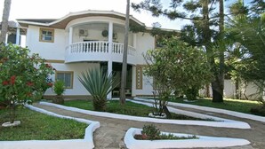 Um edifício de dois andares com varanda, rodeado por um jardim ajardinado com diversas plantas e um caminho.