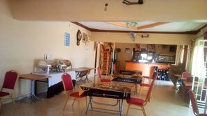 A dining area with tables and chairs, a kitchen in the background, and a ceiling fan. - The Pearl Hotel Kumi (Kumi)