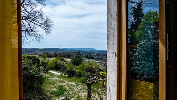 Vista desde una ventana a un paisaje rural con un muro de piedra, follaje y colinas al fondo.
