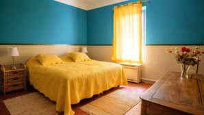 Un dormitorio con cama, mesitas de noche, escritorio, silla, ventana con cortinas y un florero con flores.