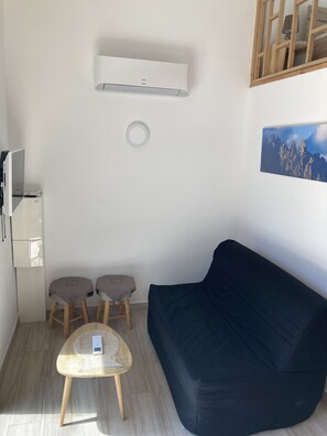Un salon moderne avec un canapé bleu foncé, une petite table ronde sur laquelle se trouve un appareil, deux tabourets recouverts de tissu, un climatiseur blanc et un tableau encadré accroché au mur.
