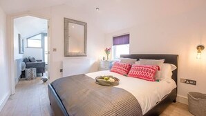 A bedroom with a large bed, a mirror, a bedside table, and a window with blinds. - Papillon, a modern Southwold flat with balcony (Southwold)