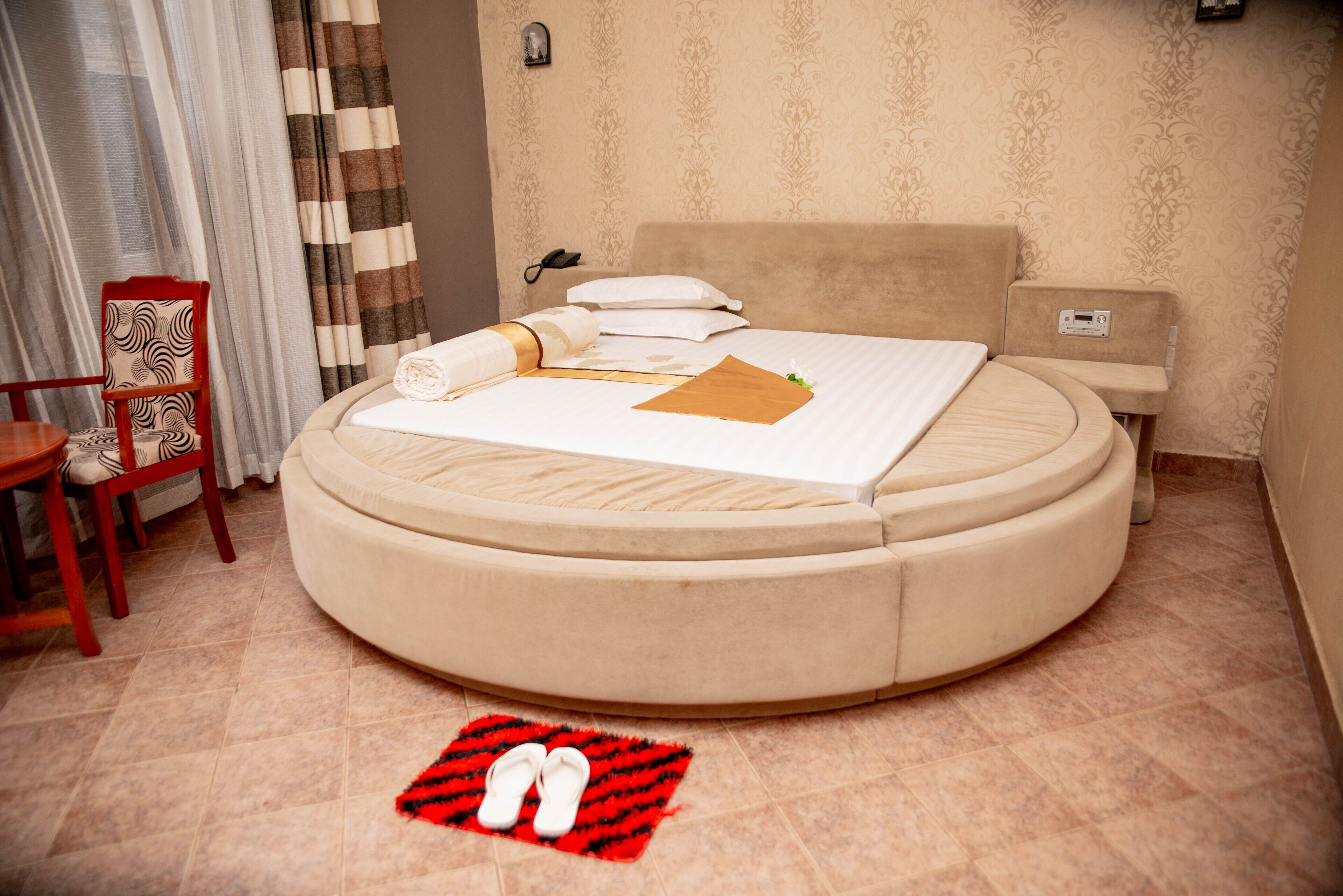 A round bed with a beige headboard, a bedside table, and a chair.