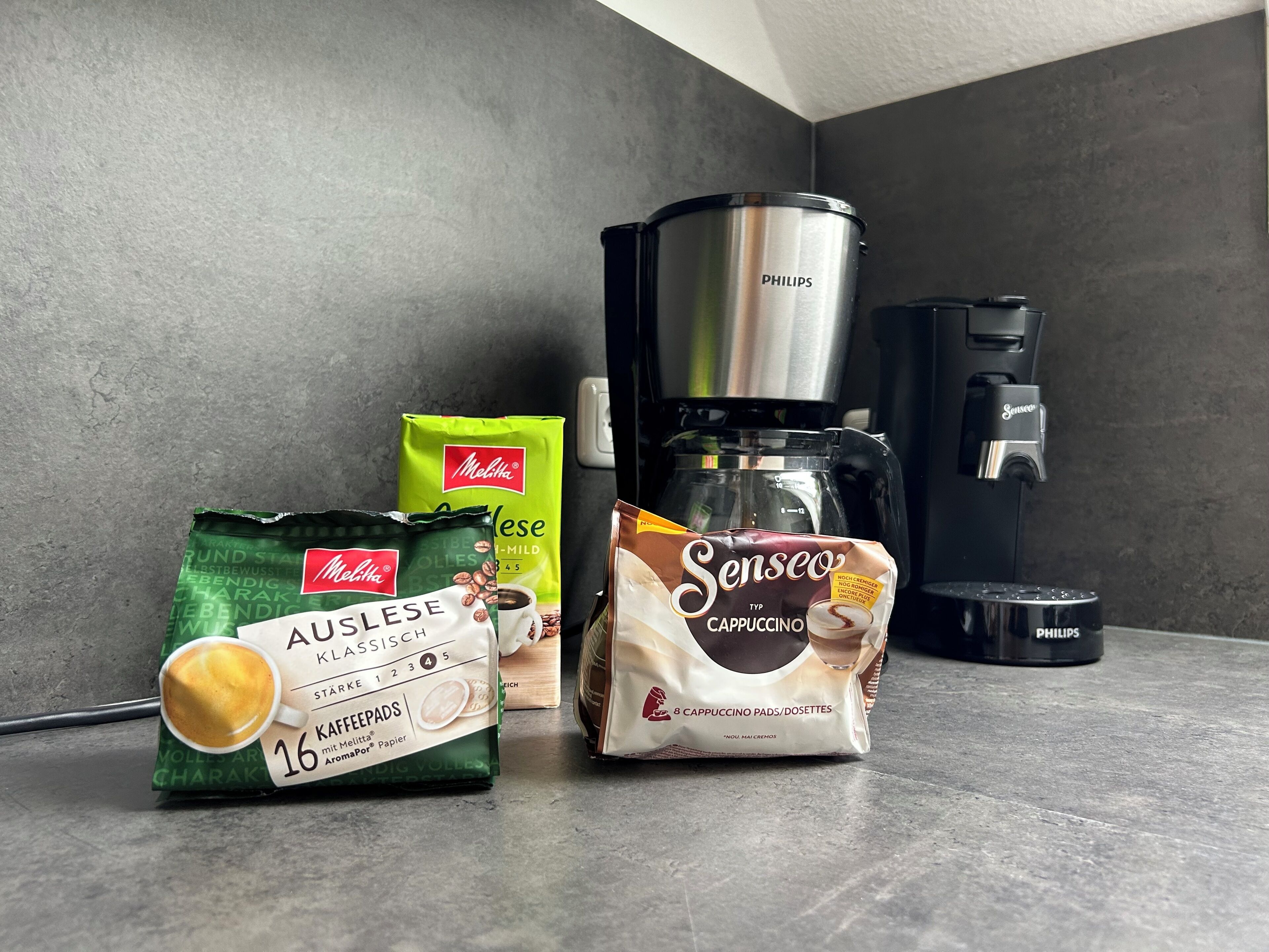 A coffee maker, a Melitta coffee pod, a Senseo cappuccino pod, and a coffee cup on a countertop.