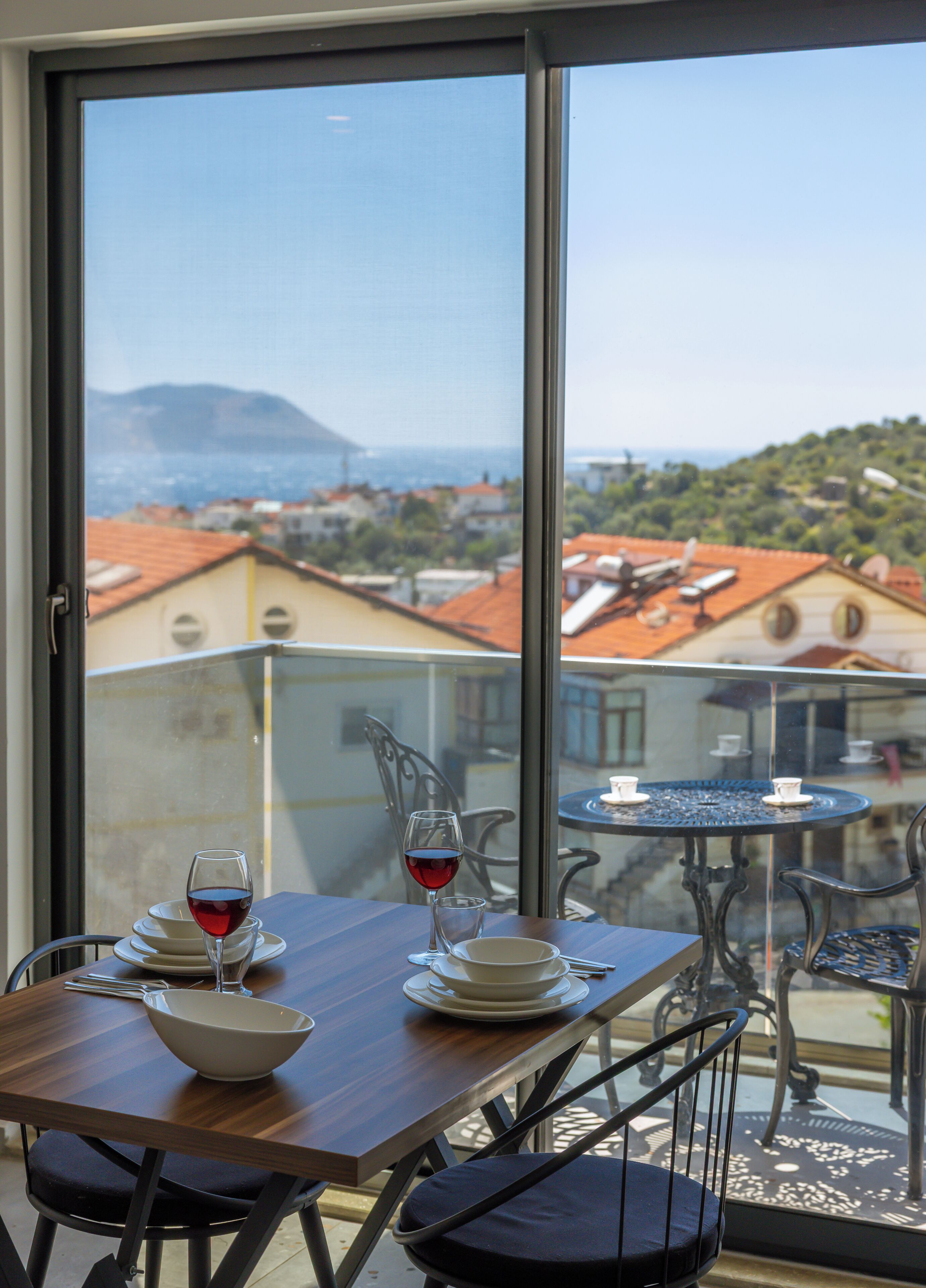 Un balcón con una mesa para dos, una copa de vino tinto y vistas a un pueblo costero.