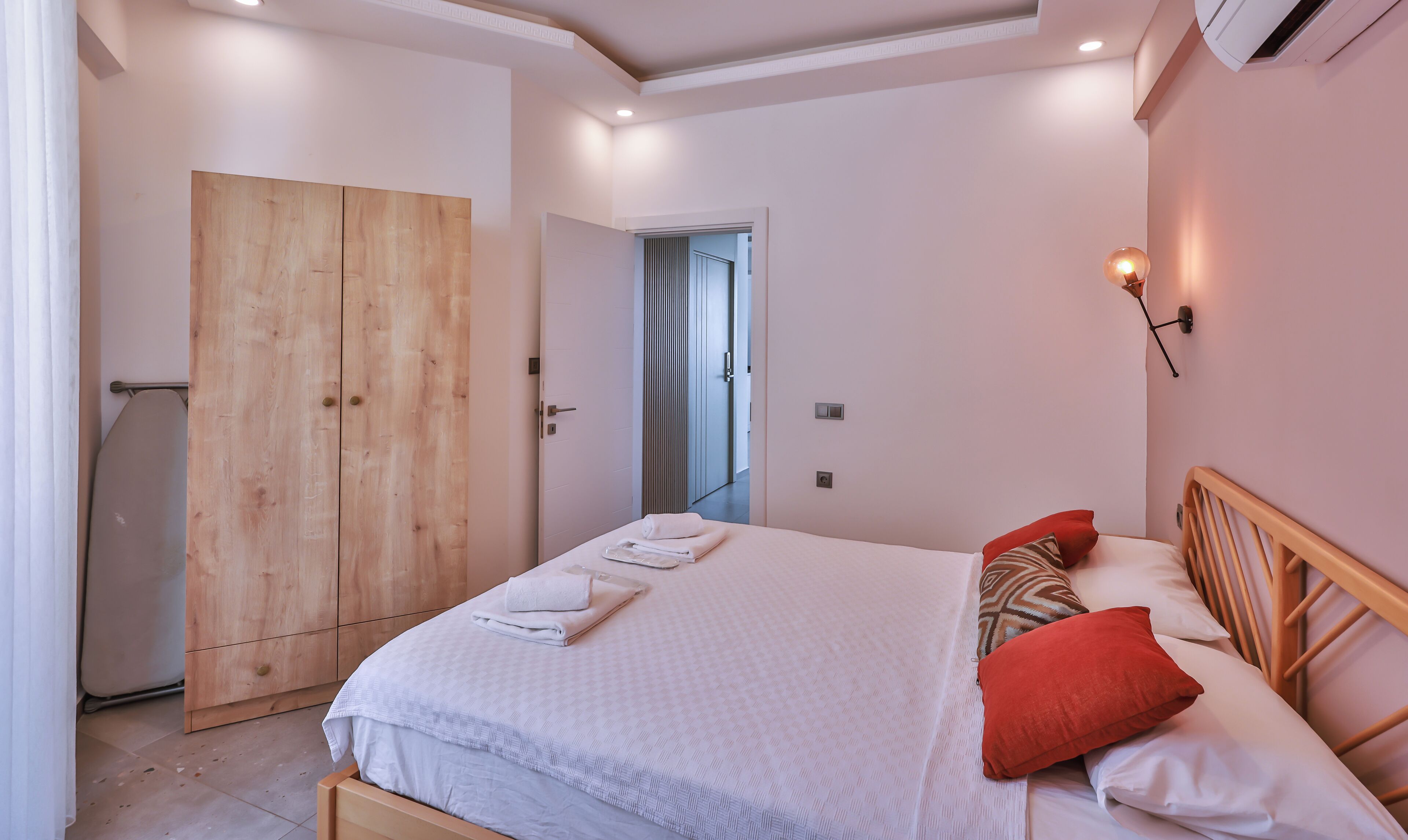 A bedroom with a wooden wardrobe, a bed with white bedding and red pillows, and a wall-mounted light fixture.
