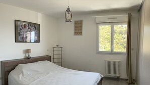 Une chambre à coucher avec un lit, un radiateur, une fenêtre avec des rideaux, une lampe fixée au mur et un tableau encadré.