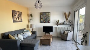 Un salon moderne avec un canapé gris, une table basse en bois et un téléviseur. Un globe terrestre est accroché au mur, ainsi qu’un tableau, et une fenêtre offre une vue sur les arbres.