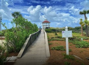 A wooden boardwalk leading to a gazebo with a red roof, surrounded by greenery and palm trees.