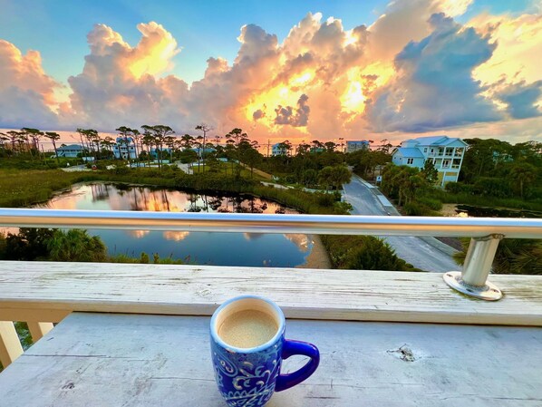 A balcony with a view of a sunset over a body of water, a road, and buildings in the distance.
