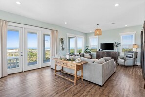 A spacious living room with a large window offering a view of the beach, a comfortable sofa, a wooden coffee table, and a flat-screen TV.