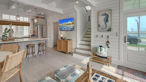 Una cocina amplia con una isla grande, un comedor con sillas de mimbre, una escalera decorada con un cuadro de caballito de mar y vistas al océano a través de una puerta de cristal.