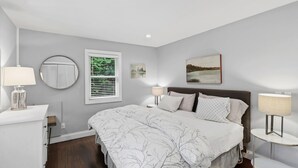 Een slaapkamer met een bed, nachtkastjes, een raam met zonwering, een ronde spiegel en een schilderij aan de muur.
