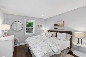 Um quarto com cama, mesas de cabeceira, uma janela com persianas, um espelho redondo e um quadro na parede.