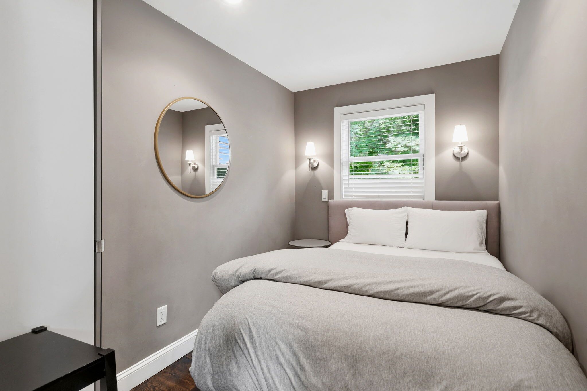 A bedroom with a bed, a round mirror, bedside lamps, a window with blinds, and a small table.