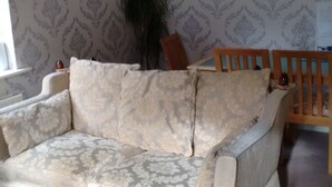 Olohuoneessa on kuviollinen tapetti, beige sohva, puinen tuoli ja huonekasvi.