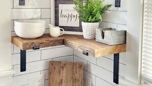 Une cuisine avec des carreaux de céramique blancs disposés en motif métro, des étagères en bois, une passoire suspendue, un bocal Mason, un panneau encadré, une plante en pot et une planche à découper en bois.