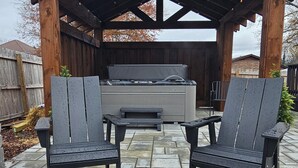 Une terrasse dotée d’un foyer extérieur, de deux fauteuils Adirondack et d’un jacuzzi, le tout installé sous une pergola en bois.