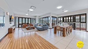 A spacious living area with a wooden floor, a blue sofa, a wooden dining table with chairs, and large windows offering a view of the outdoors.