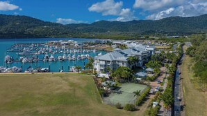 A marina with boats, a large building with a tennis court, and a hilly landscape in the background.