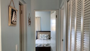 Un couloir menant à une chambre à coucher, qui contient un lit, un luminaire au plafond et une porte avec des stores.