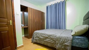 A bedroom with a bed, a wardrobe, and a bathroom visible through an open door. - Cozy Family-sized 2 Bedroom Apartment with stable Wi-fi & conducive work station (Ruiru)